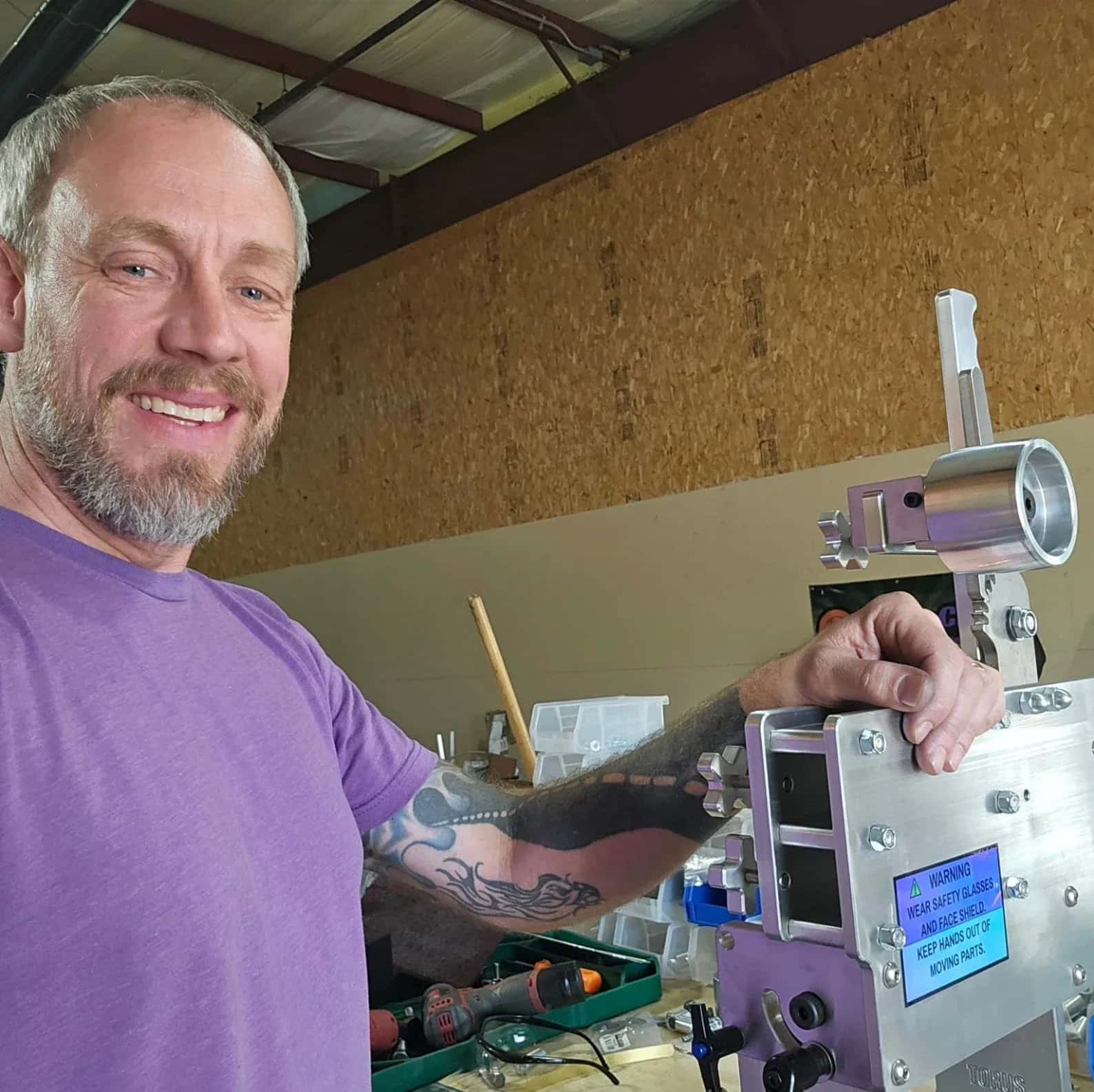 Smiling machinist resting his tattooed arm on a Torus CNC belt grinder frame in the workshop