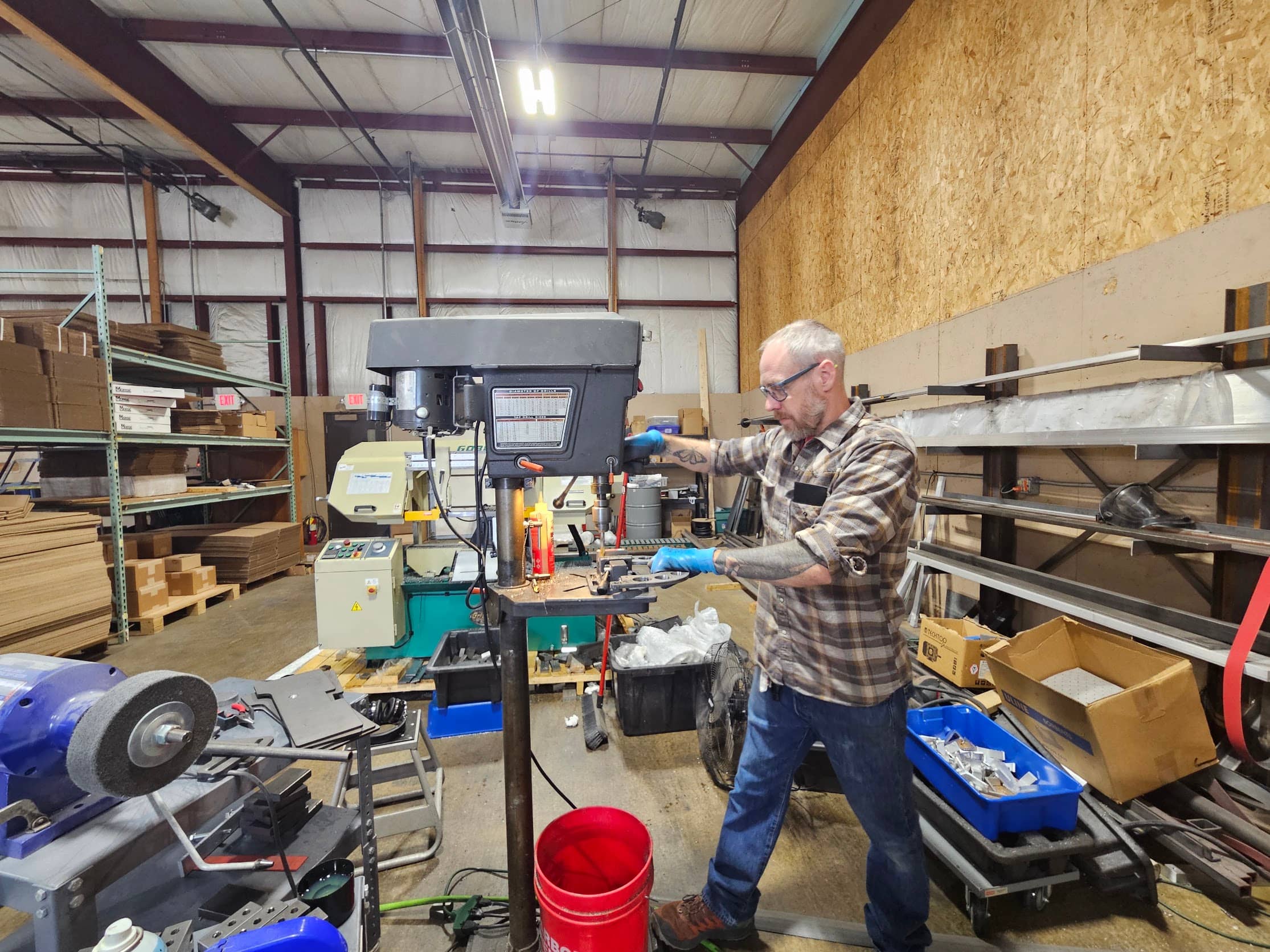 Rich Linville in safety glasses running a grinder part on the drill press, steel stock racked behind him