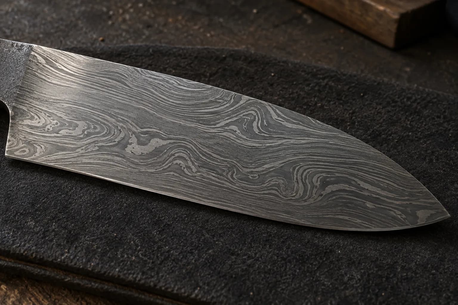 Etched Damascus blade lying flat on dark felt, fine silver layer lines swirling against darker carbon steel.