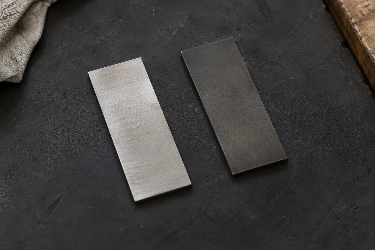 Two plain steel coupons side by side on a dark mat, the left bright silver-gray and the right dark etched gray.