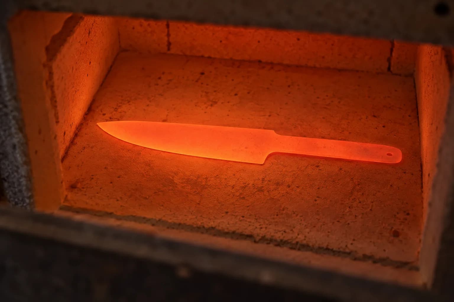 Evenly glowing orange blade blank with a tang hole lying flat on the floor of an electric kiln chamber.