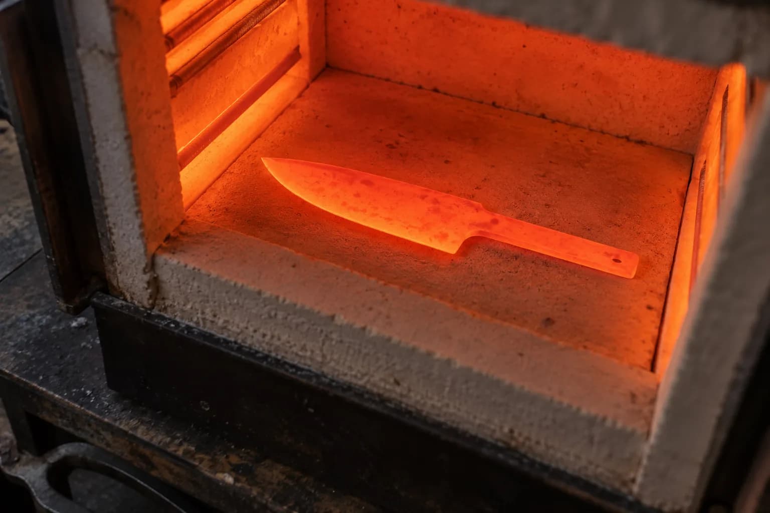 Plain knife blade with tang glowing evenly orange-red on the ceramic floor of an electric heat-treat kiln.