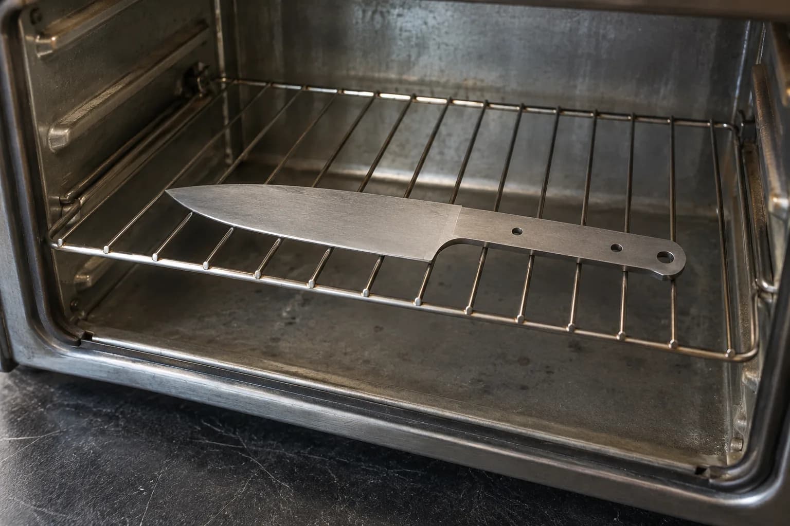 Unhandled steel knife blank lying flat on the wire rack inside an empty stainless steel oven.