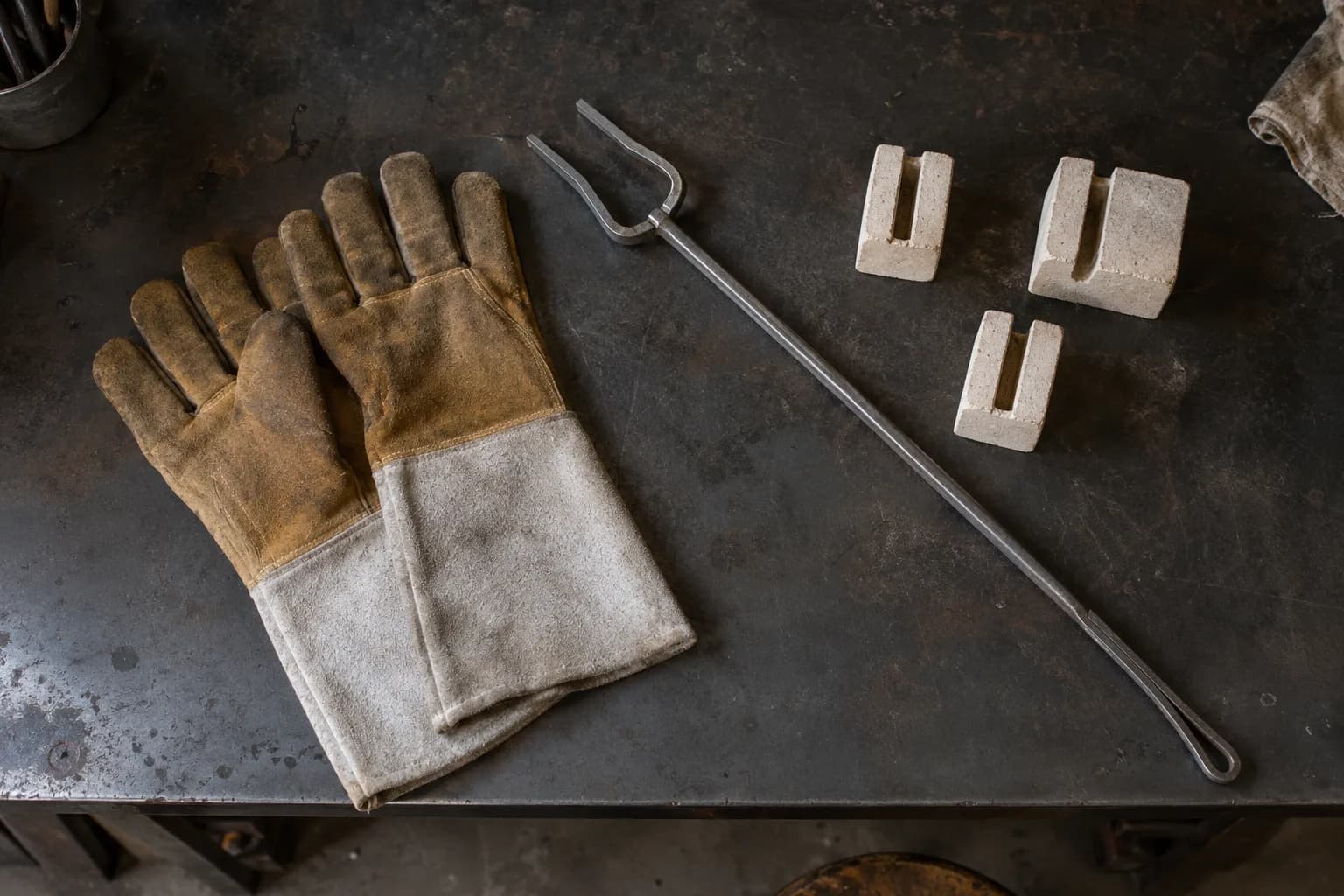Overhead view of leather heat gloves, a long steel handling fork and three slotted ceramic blade holders on a steel bench.