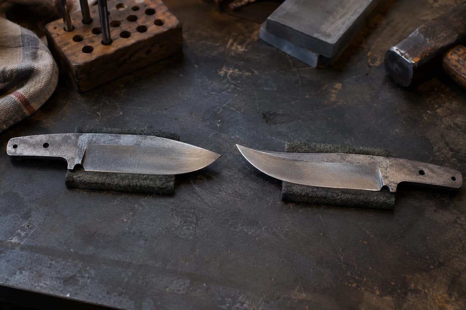 Drop-point and trailing-point knife blanks on separate felt pads, tips facing each other on a dark steel bench.