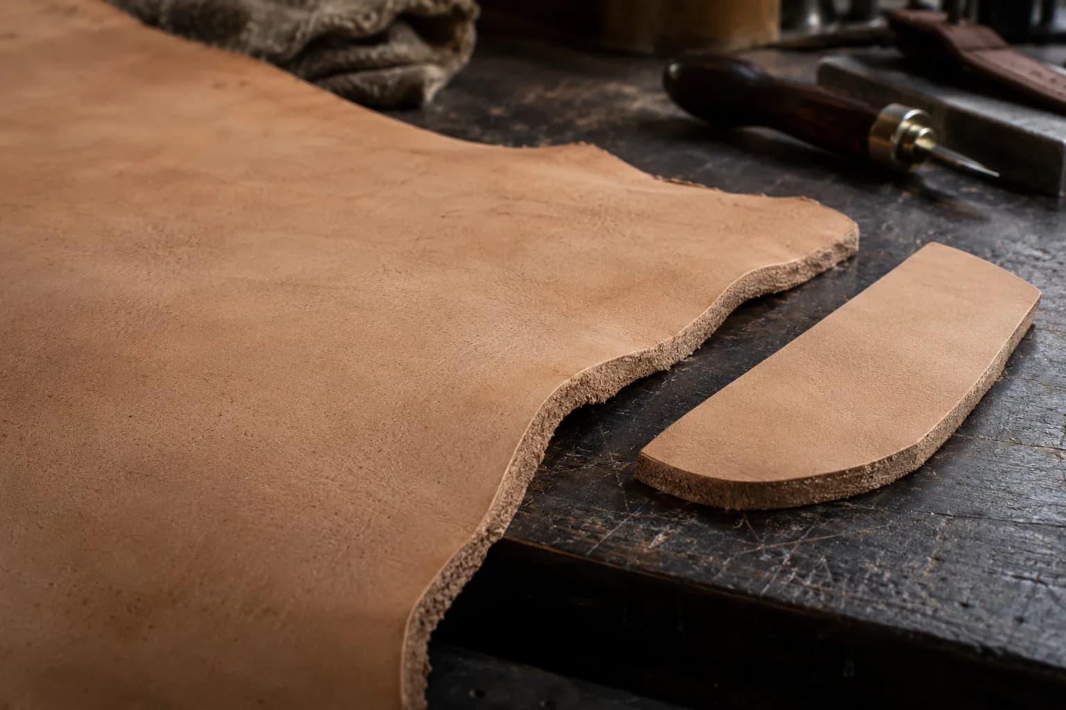 Thick undyed vegetable-tanned leather hide with a raw cut edge beside a flat sheath-shaped offcut on a dark bench
