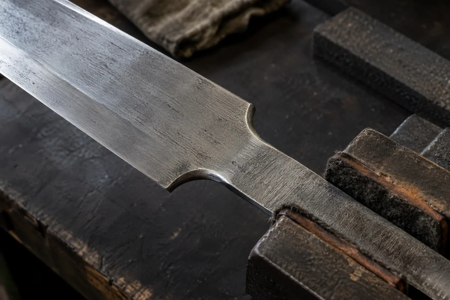 Close view of a bare sword blade's radiused shoulders and flat tang held in felt-padded wooden jaws on a dark bench