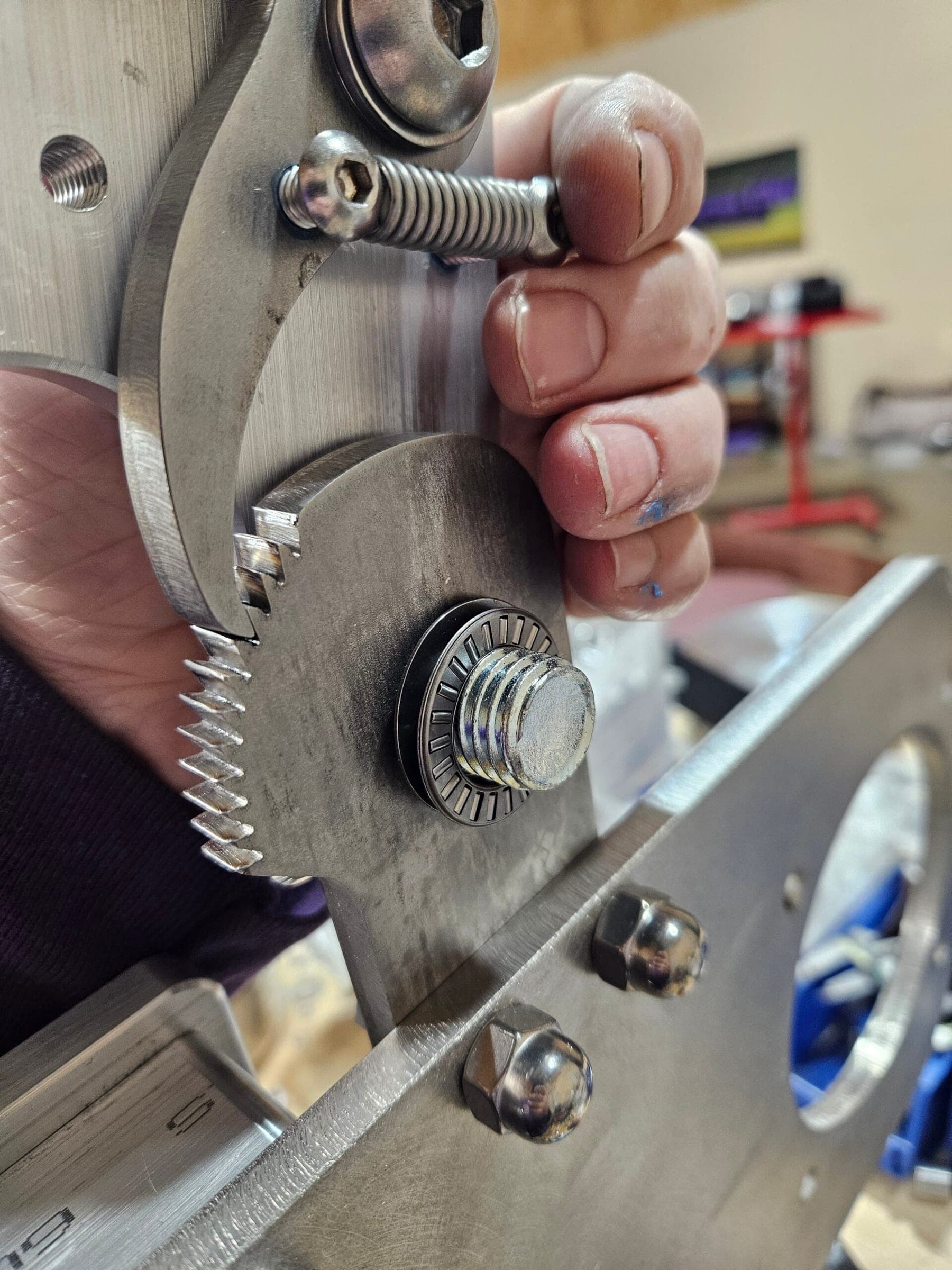 Hand gripping a stainless ratchet tension arm, with teeth, pivot bolt, and spring visible.