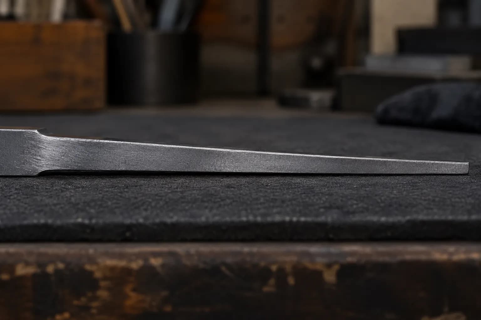 Side view of a knife blank's spine on a dark pad, thick at the ricasso and thinning to a blunt tip
