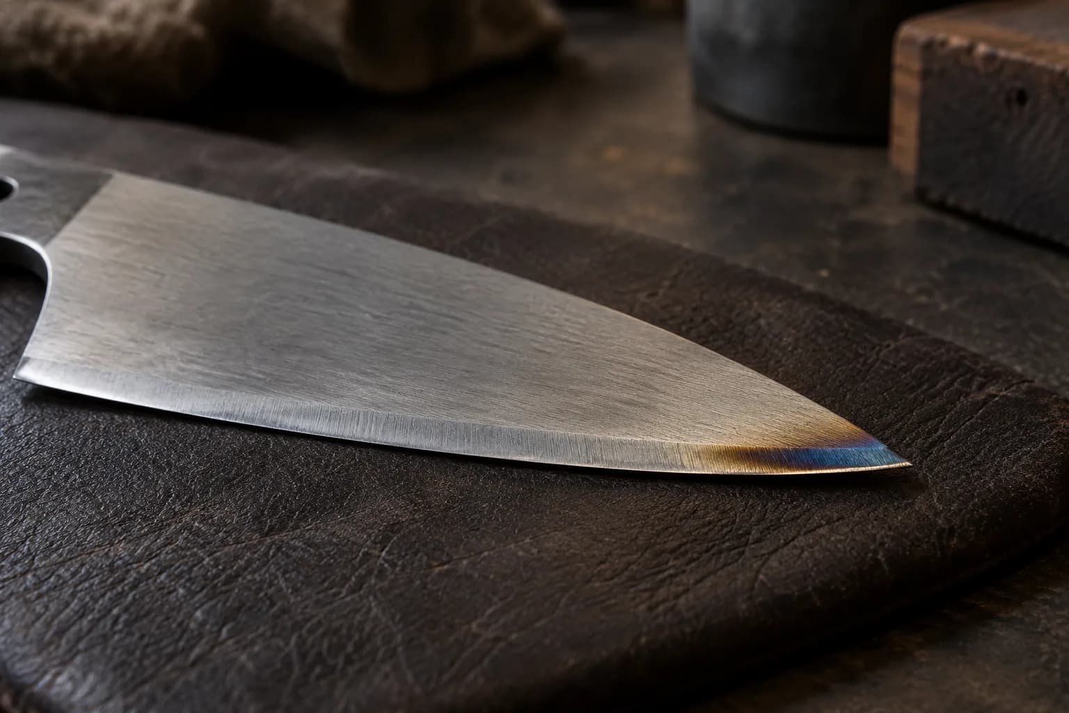 Satin knife blade on a dark leather pad with a straw-to-blue heat tint along the edge near the tip