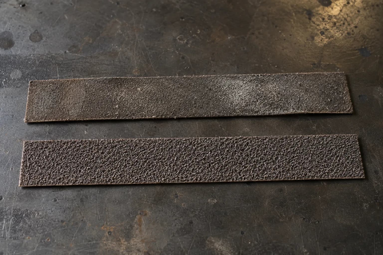 Two cut belt strips on a steel bench, a gray glazed worn one above a fresh strip with open sharp grain