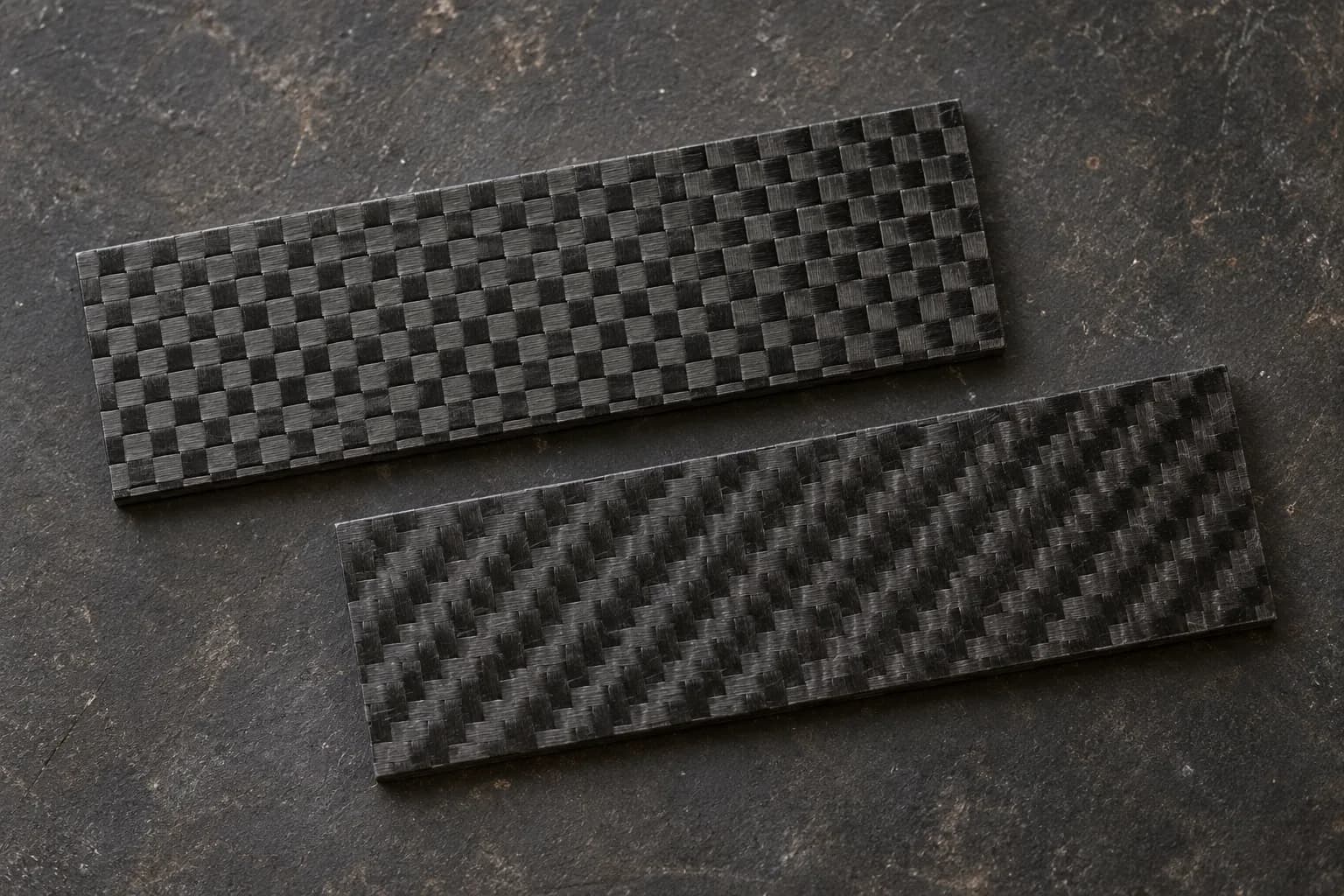 Two flat black carbon-fiber handle blanks on a dark surface, one plain checkerboard weave and one diagonal twill.