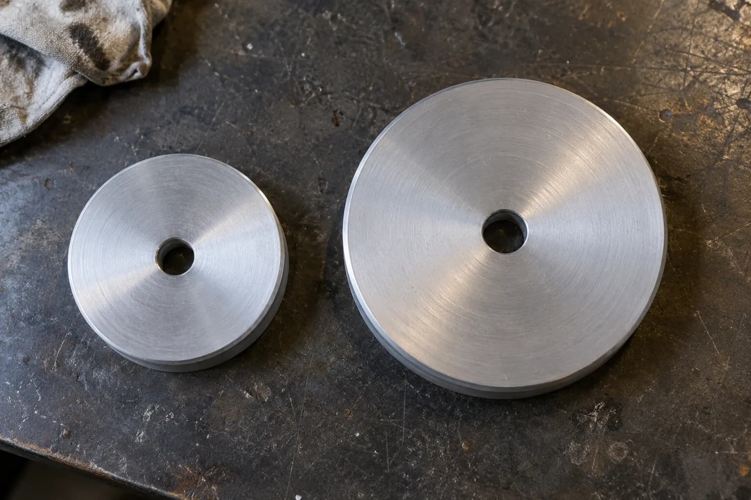 Two plain brushed aluminum drive wheels, one smaller and one larger, lying flat with center bores on a dark steel bench.