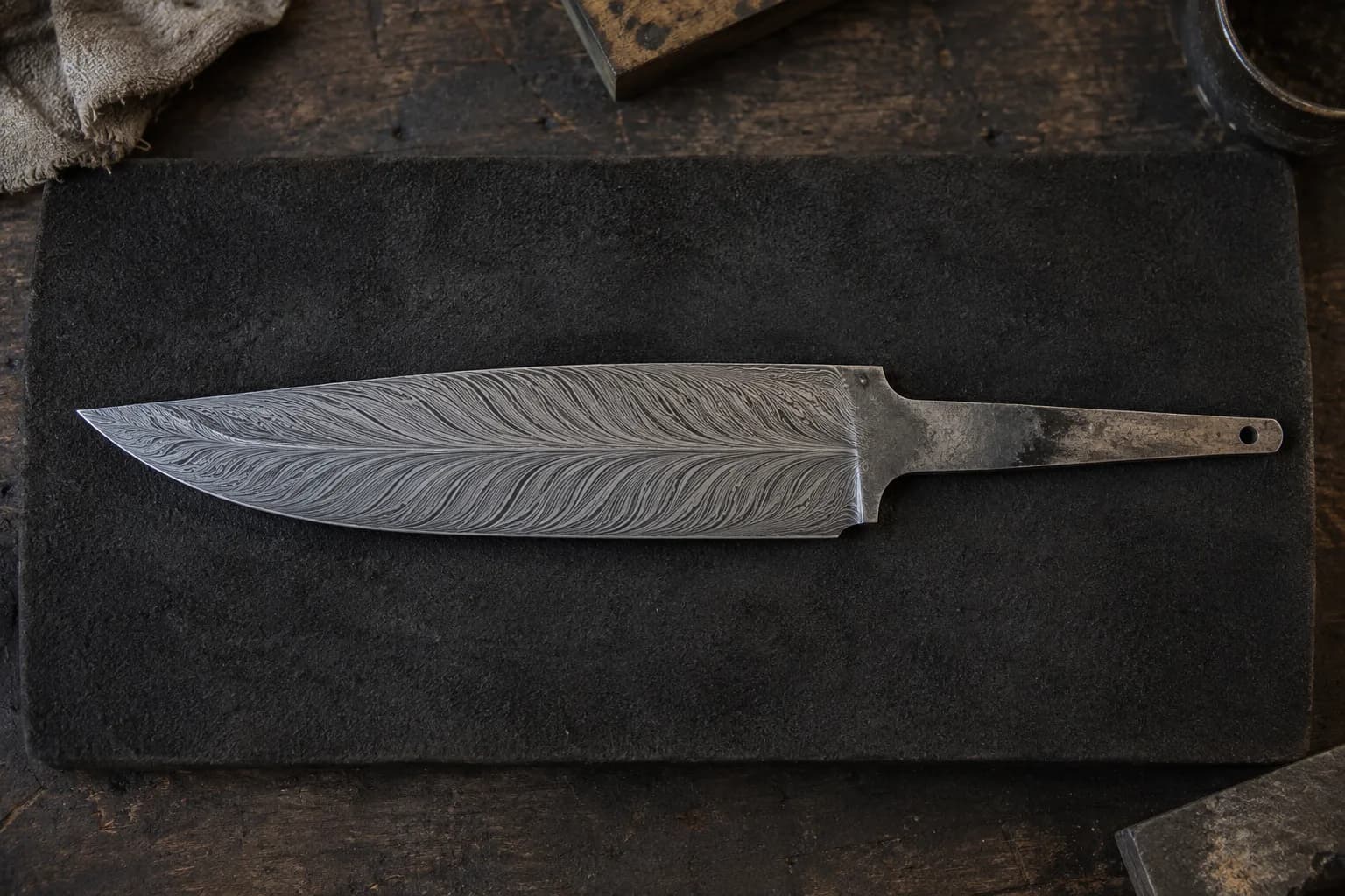 Overhead view of an etched feather-Damascus blade on a charcoal pad, barbs sweeping from a central quill.