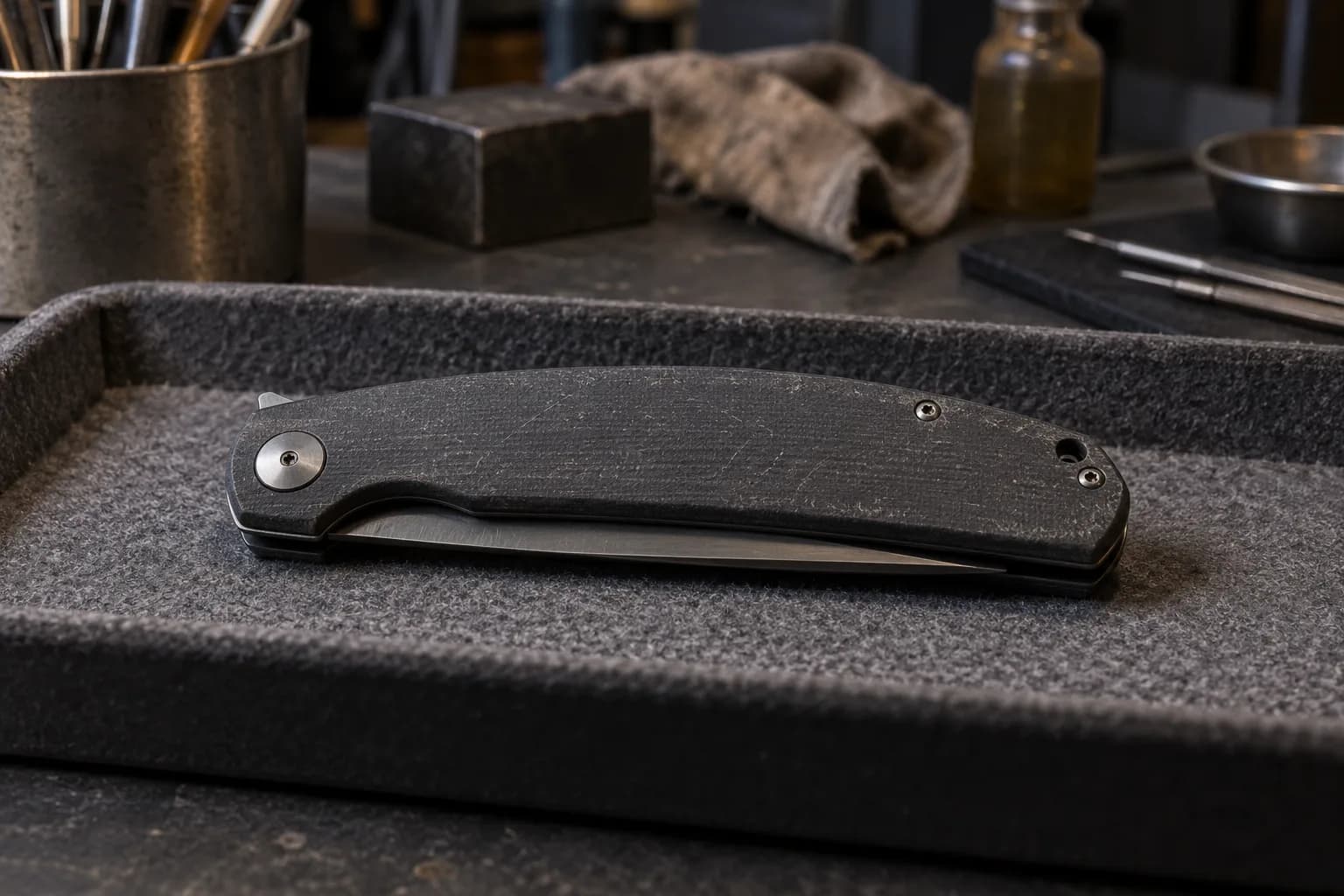 Closed black-handled folding knife on a gray felt tray with a thin sliver of blade showing below the handle, slightly ajar.