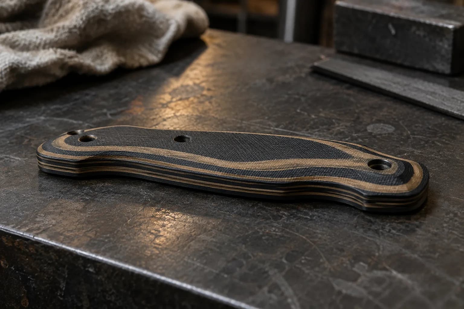 Black and tan layered G10 handle scale with four pin holes, its contoured face exposing tan bands, on a dark steel bench