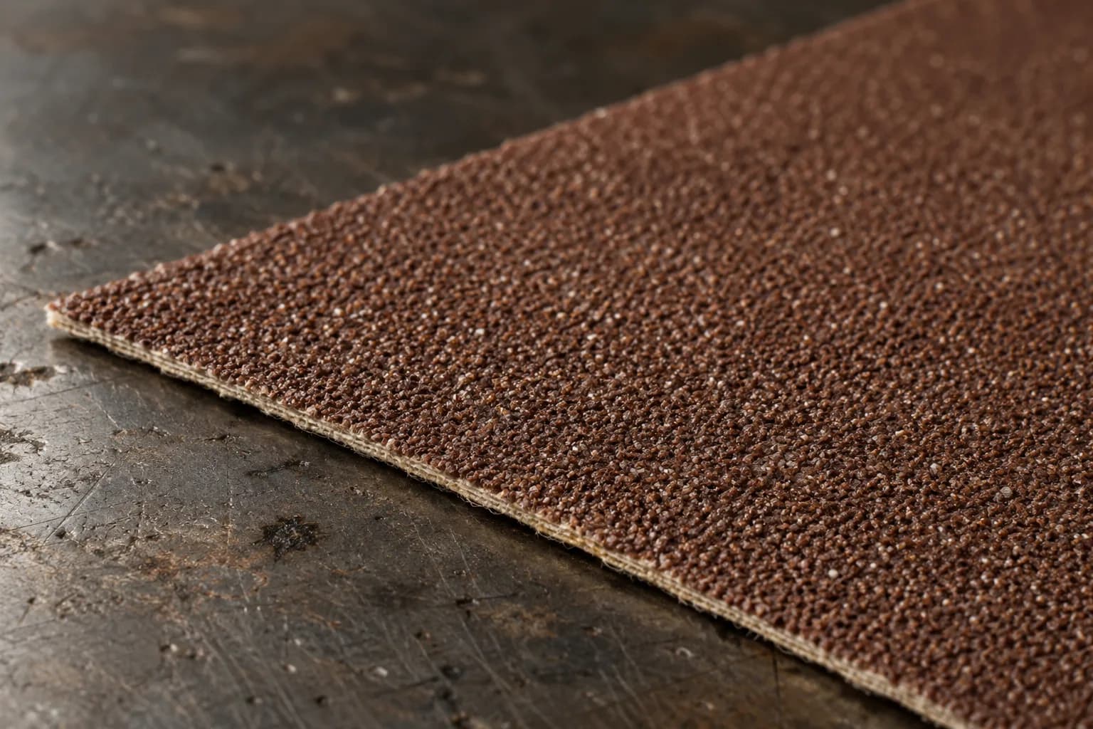 Close view of a brown aluminum-oxide belt strip on a scarred steel bench, grit surface and cloth backing edge visible.