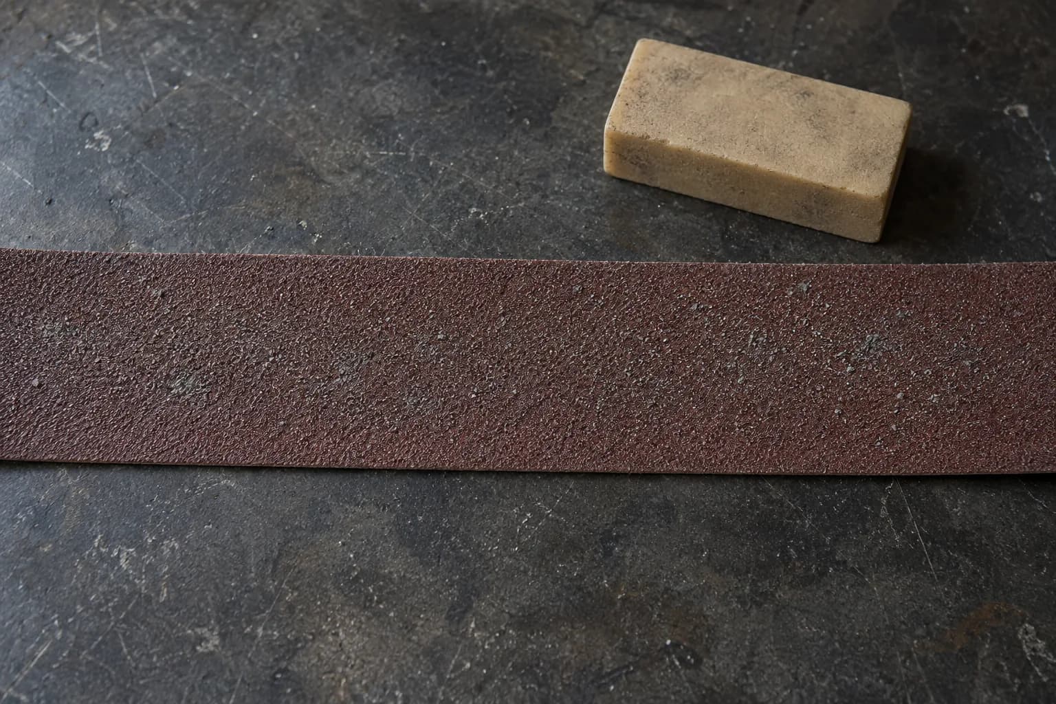 Red-brown abrasive belt laid flat with light gray swarf in the grit, beside a dirty tan rubber belt eraser on a dark bench.