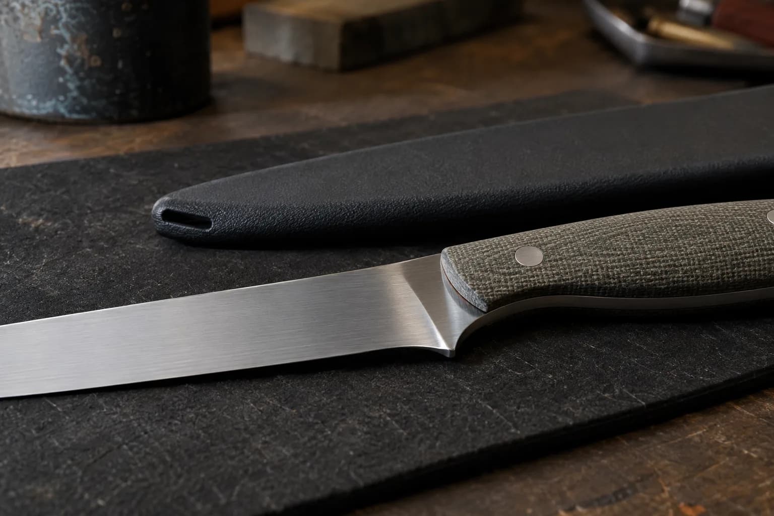 Satin-finished fillet knife with green canvas micarta handle on a dark pad beside a black molded sheath with a drain slot at its tip