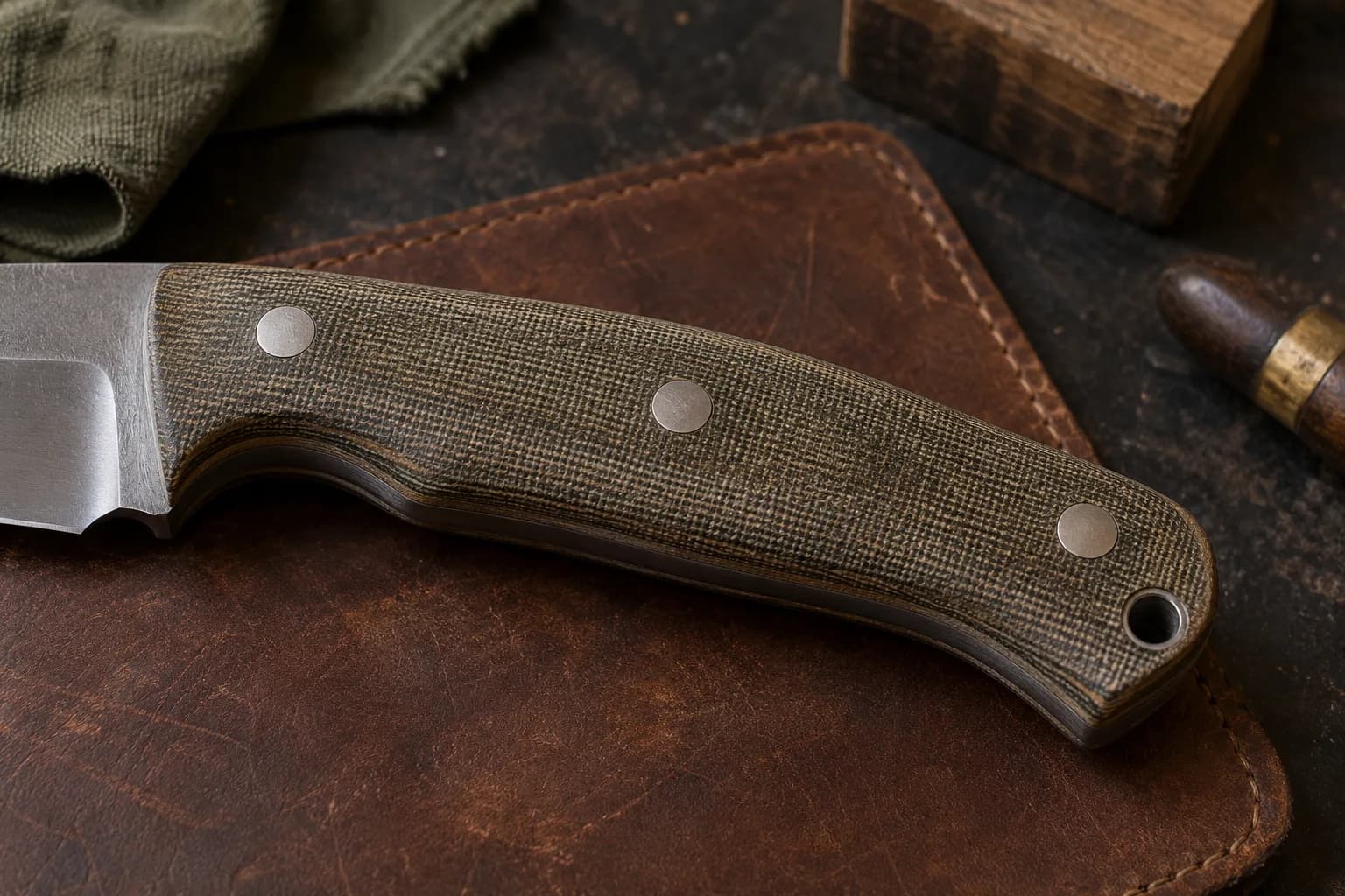 Canvas Micarta knife handle with three flush pins and layered edges on a worn brown leather pad, blade at left.