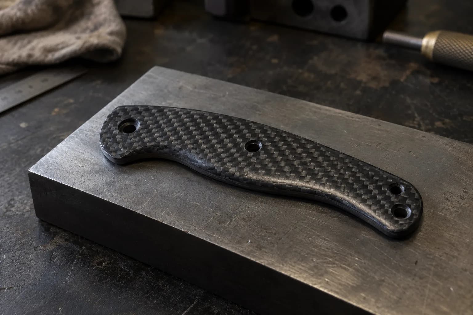 Shaped black carbon-fiber handle scale with a woven twill face and four mounting holes resting on a steel block.