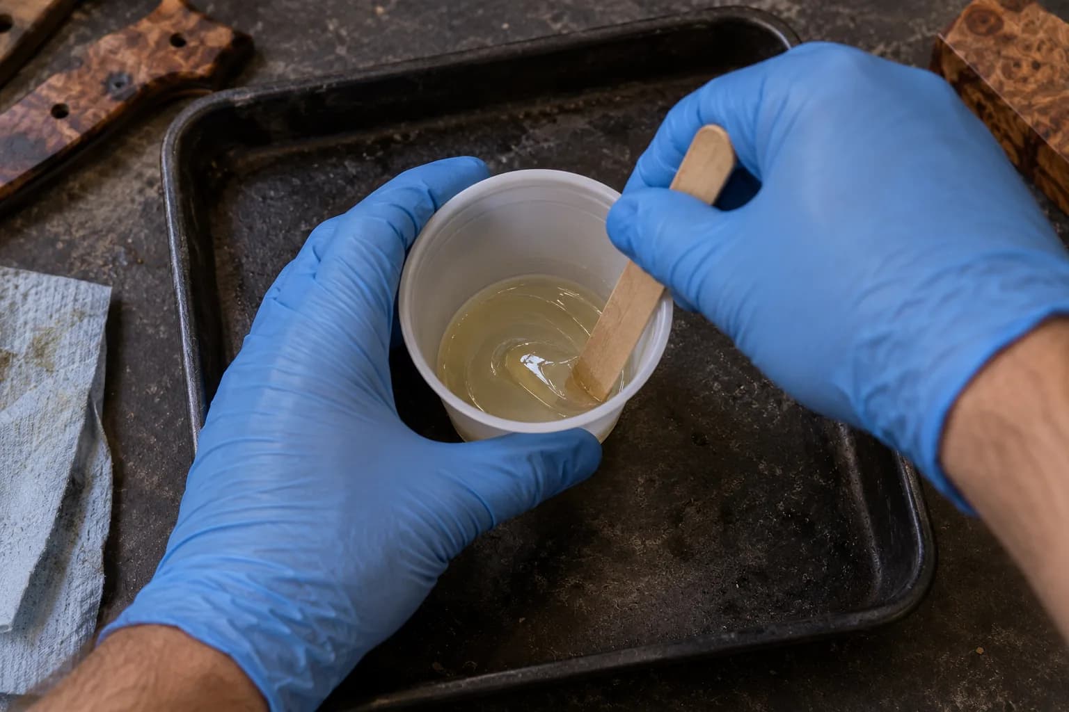Blue-gloved hands holding a clear cup of mixed epoxy while a wood stick scrapes along the cup wall.