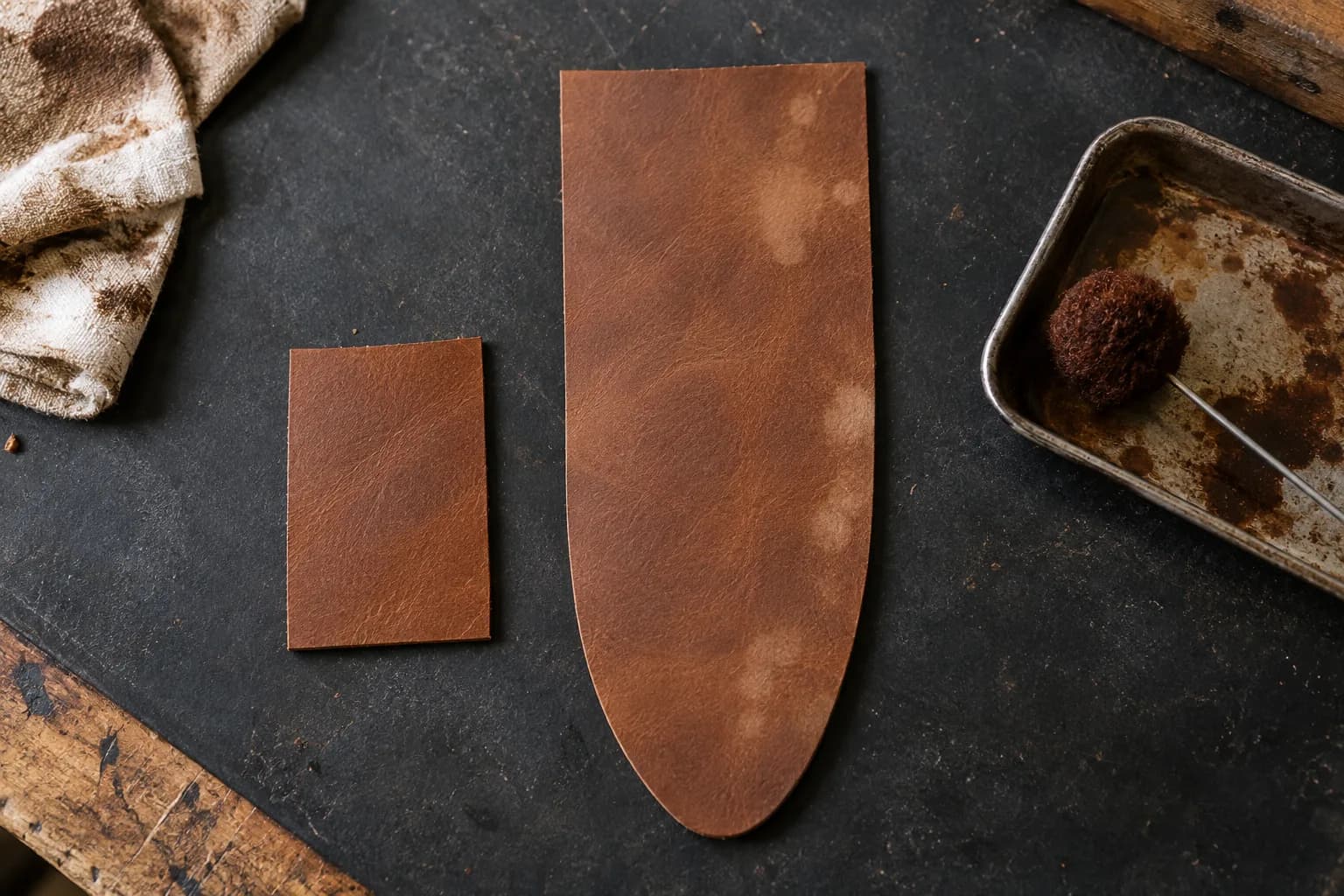 Top-down view of a small evenly dyed leather scrap beside a flat sheath panel with pale blotches, a dauber in a tray nearby.
