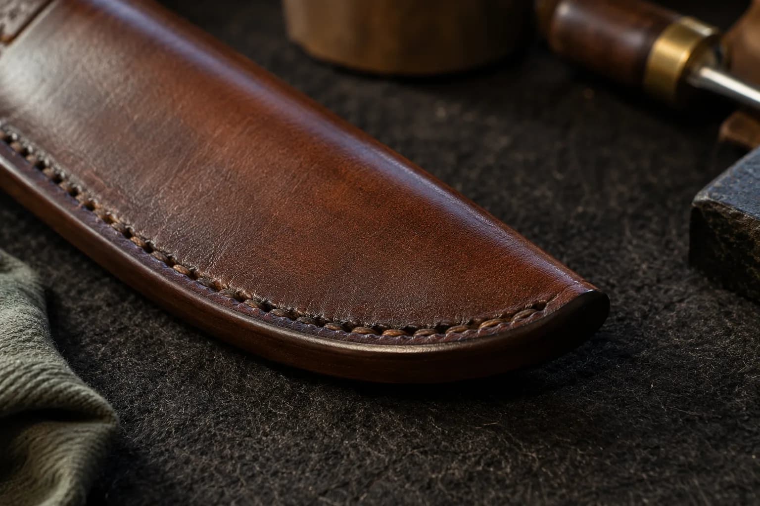 Close view of a dark burnished, flush edge and hand stitching at the tip of a finished brown leather sheath on dark felt.