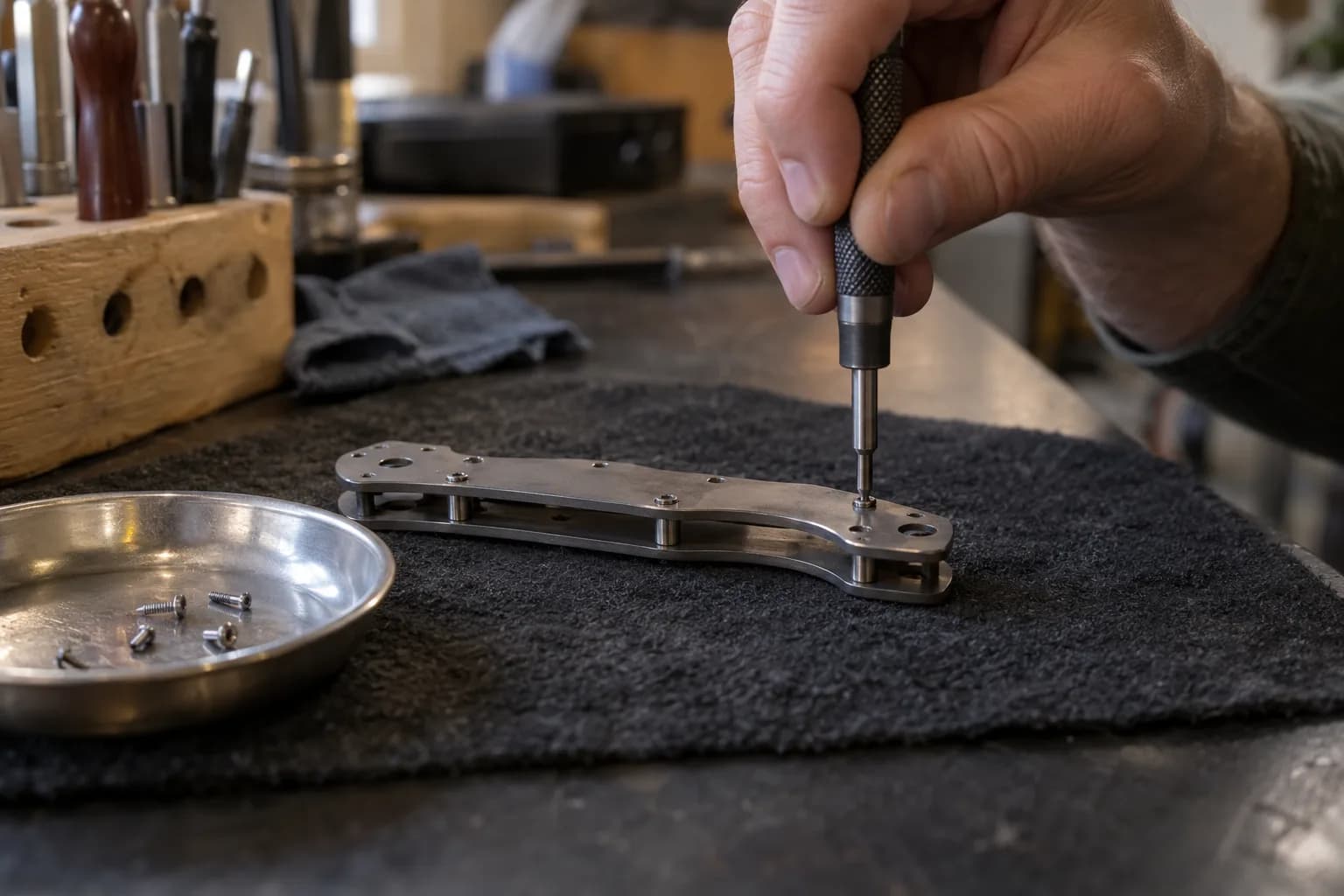 Hand holds a knurled driver square over a body screw on a folder chassis with standoffs; spare screws sit in a dish.