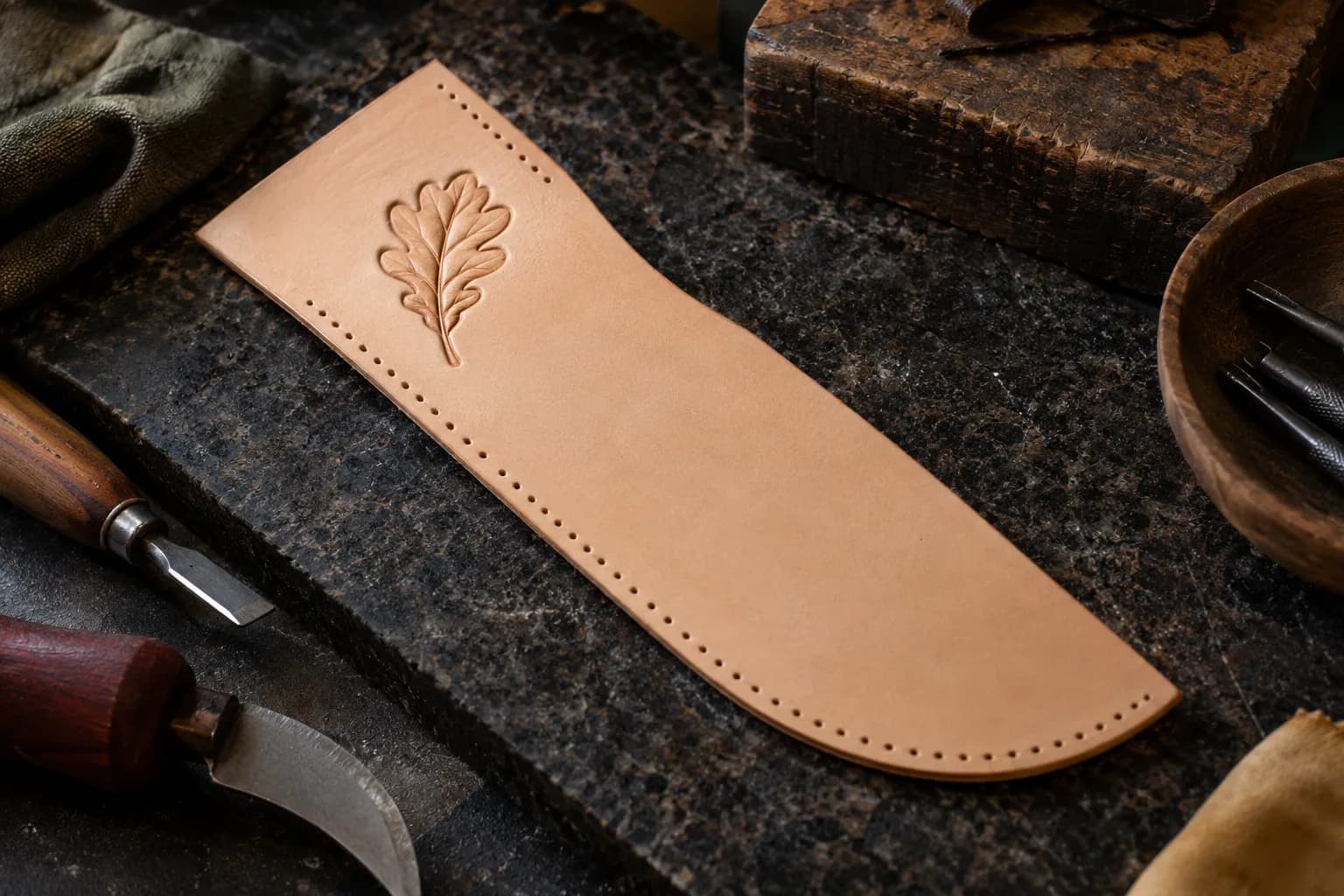 Flat tan leather sheath panel with one small carved oak leaf and plain untooled leather, on dark granite beside leather tools.