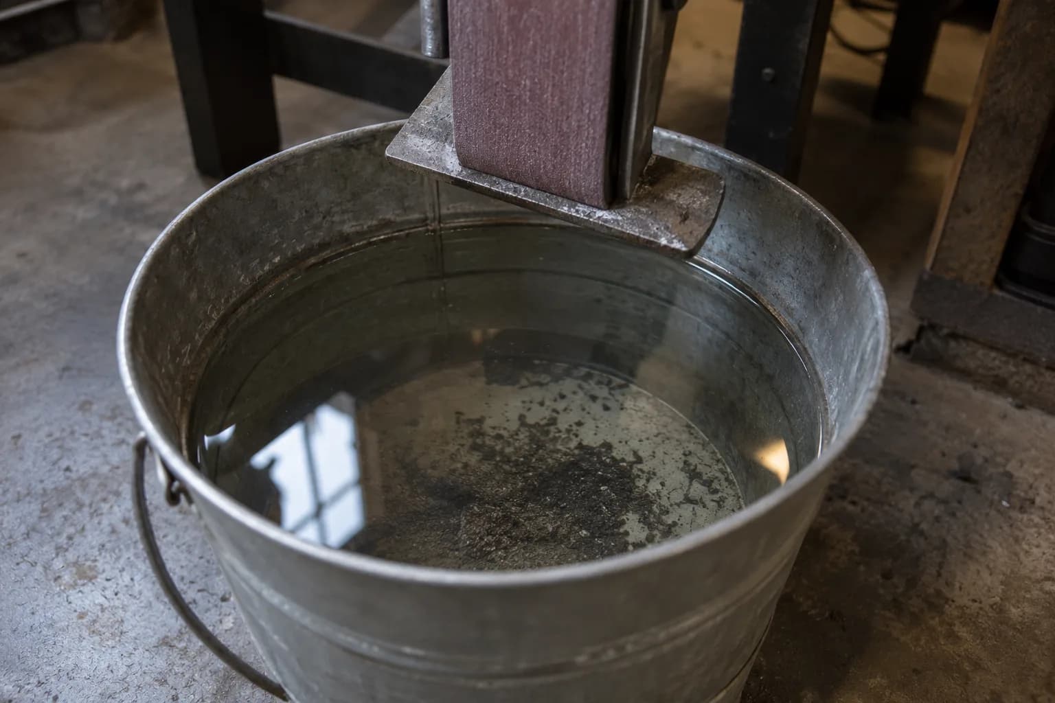 Galvanized bucket of water with dark grit on its bottom, set under the work rest of a stationary abrasive belt.
