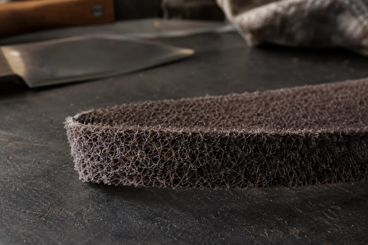 Close view of a dark nonwoven conditioning belt loop on a charcoal bench, open fibers visible, knife blade blurred behind