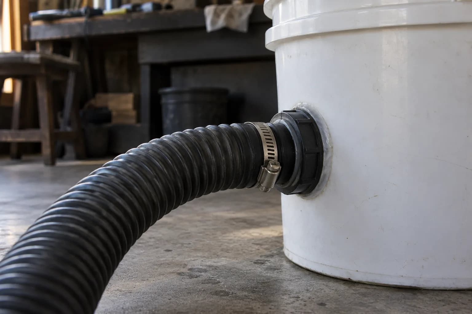 Corrugated black hose clamped to a fitting sealed through the side of a white bucket, on a concrete shop floor
