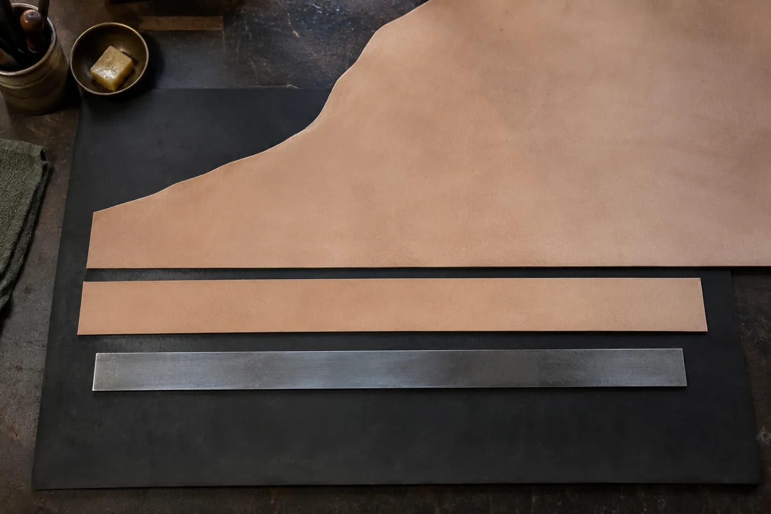 Tan leather strip cut straight from a hide on a dark mat, with a plain unmarked steel straightedge laid below it.