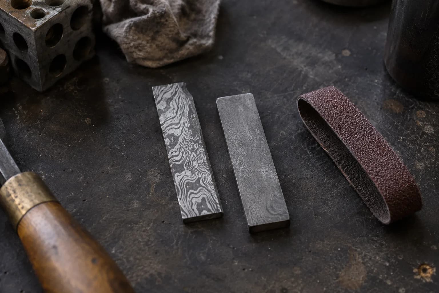Two small etched Damascus test coupons, one bold and one flat gray, beside a brown sanding belt loop on a dark bench
