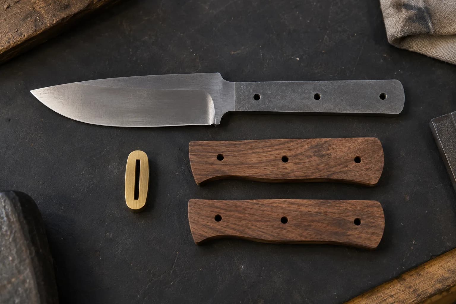 Knife blank with three pin holes beside two walnut handle scales and a slotted brass guard on a dark slab.