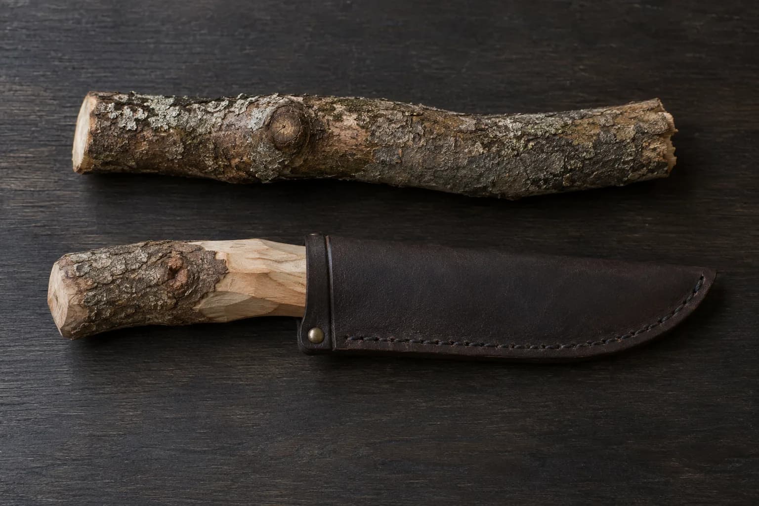 Bark-covered branch segment with a knot above a partly carved branch-handled knife in a plain brown leather sheath on a dark bench.