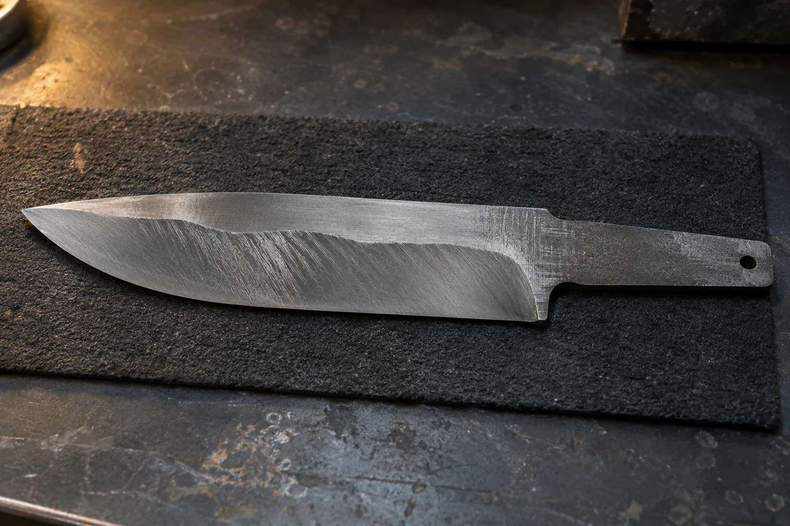 Unfinished knife blank on a black felt pad with a wavy, uneven grind shoulder and patchy coarse scratches.