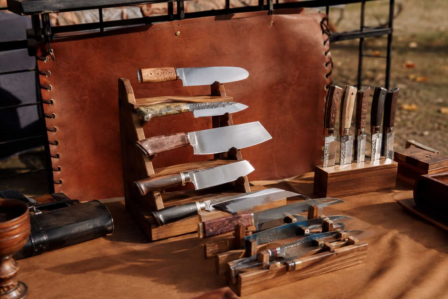 Handmade chef and kitchen knives displayed on a wooden stand at an outdoor craft-show table with a leather banner