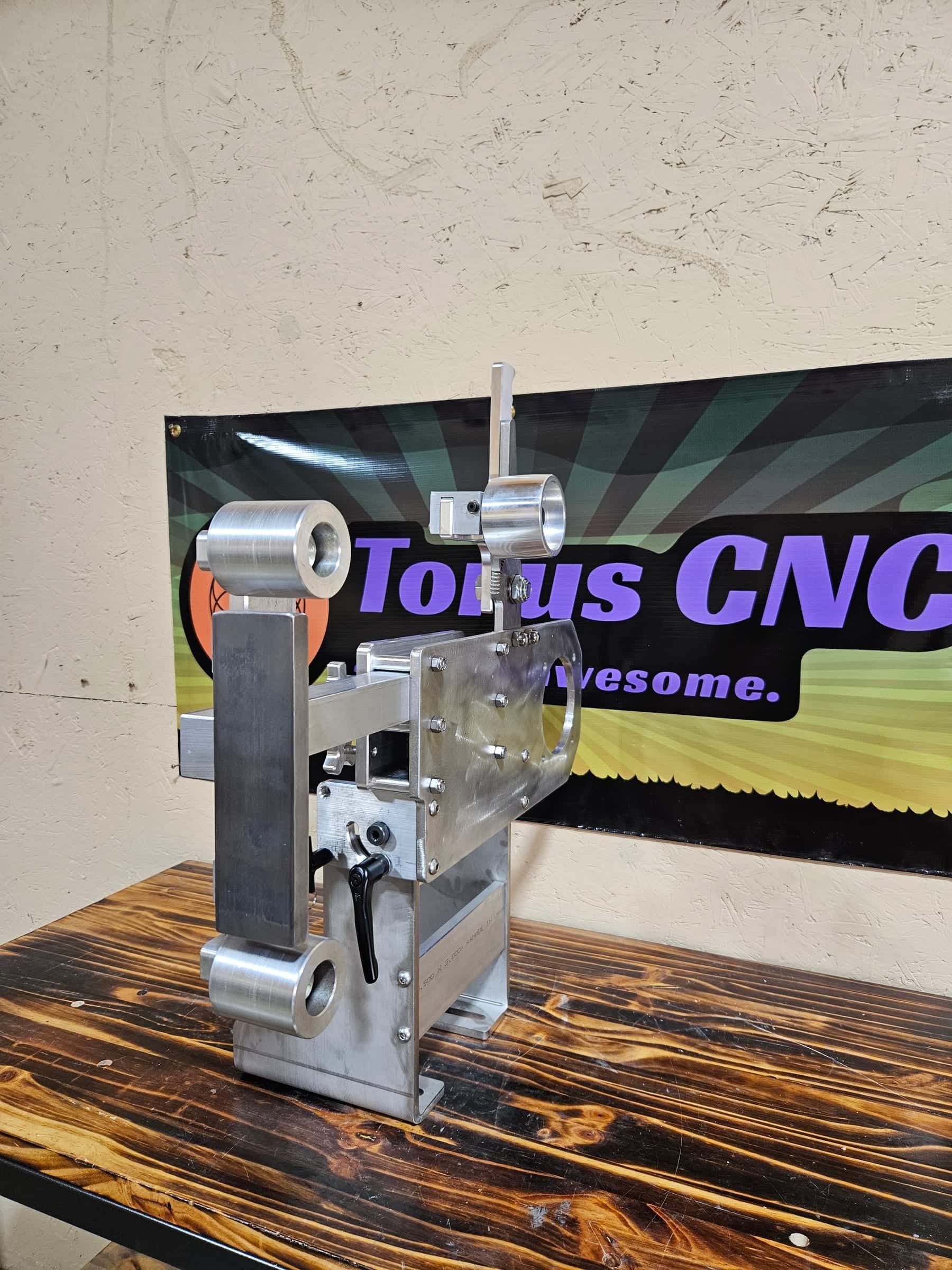 Aluminum Torus CNC belt grinder tracking arm with idler wheels standing on a workbench in front of the shop banner