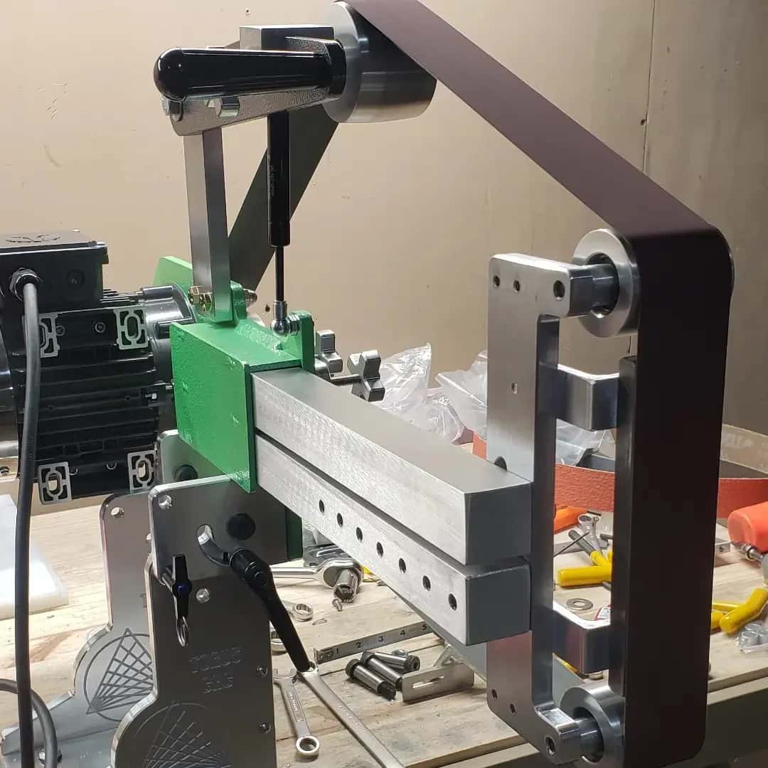 Close-up of a Torus CNC belt grinder's tooling arm, tension knob, and drive wheel assembly on a bench