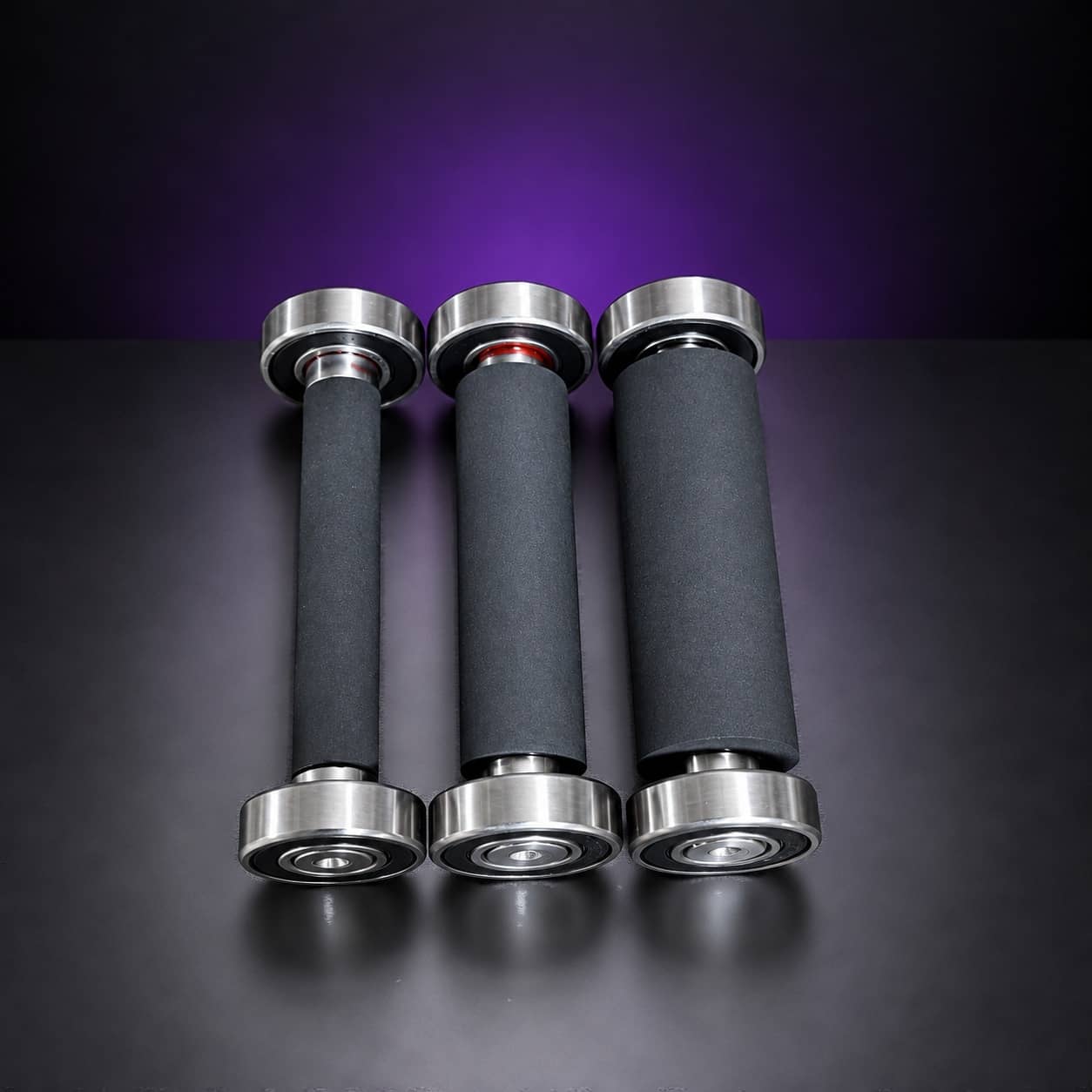 Three black rubber small wheels on bearings, from narrow to wide, standing in a row, on black with a purple glow