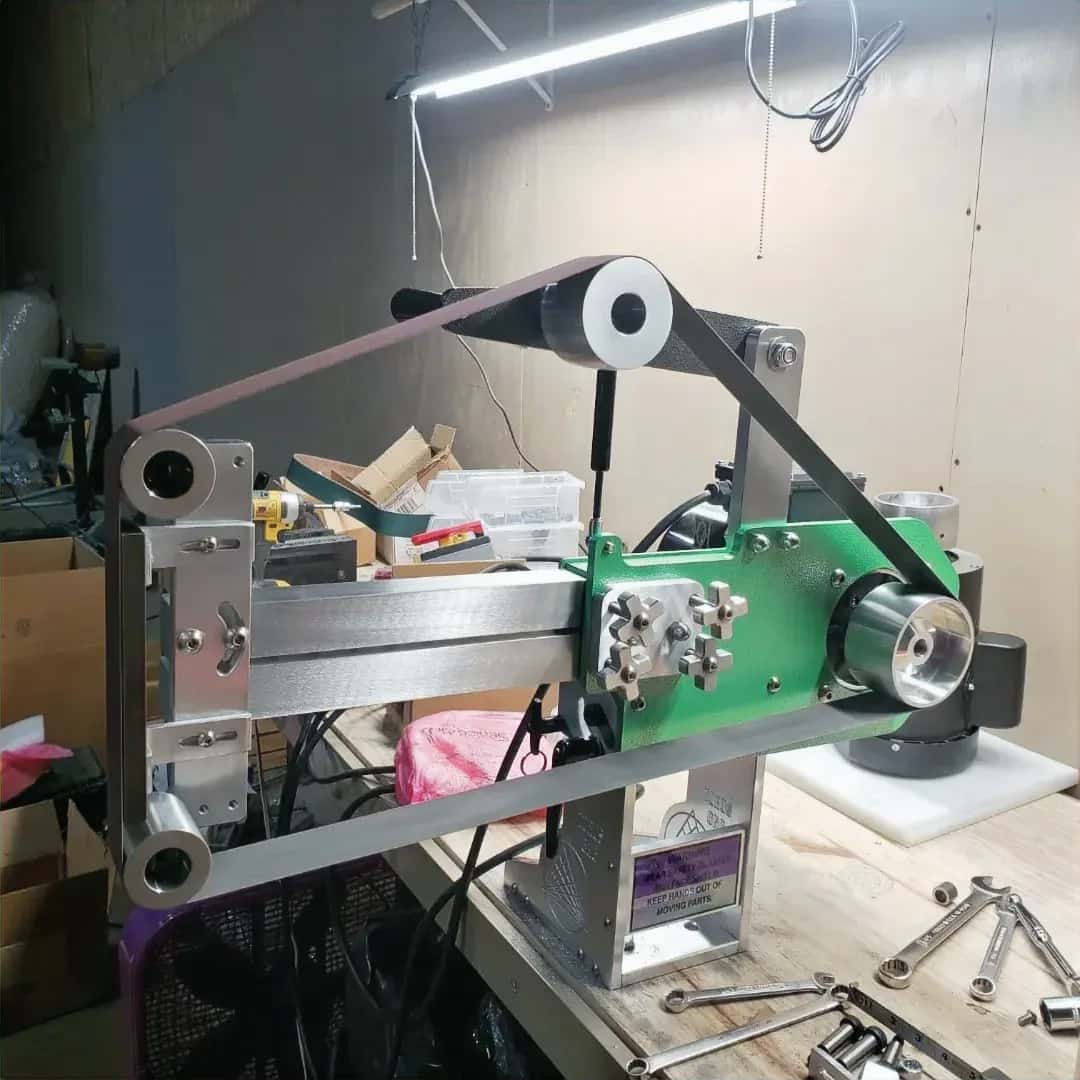 Belt grinder assembly mounted on a workbench, with a sanding belt, tool boxes, and shop clutter nearby