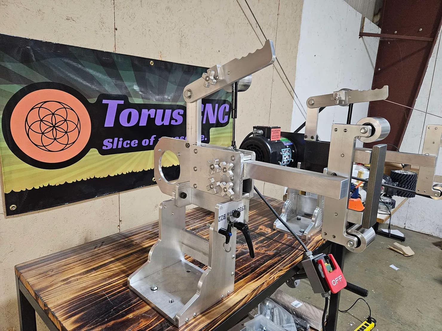 Two Torus CNC tooling arm assemblies with drive wheels set up on a workbench beside the shop banner