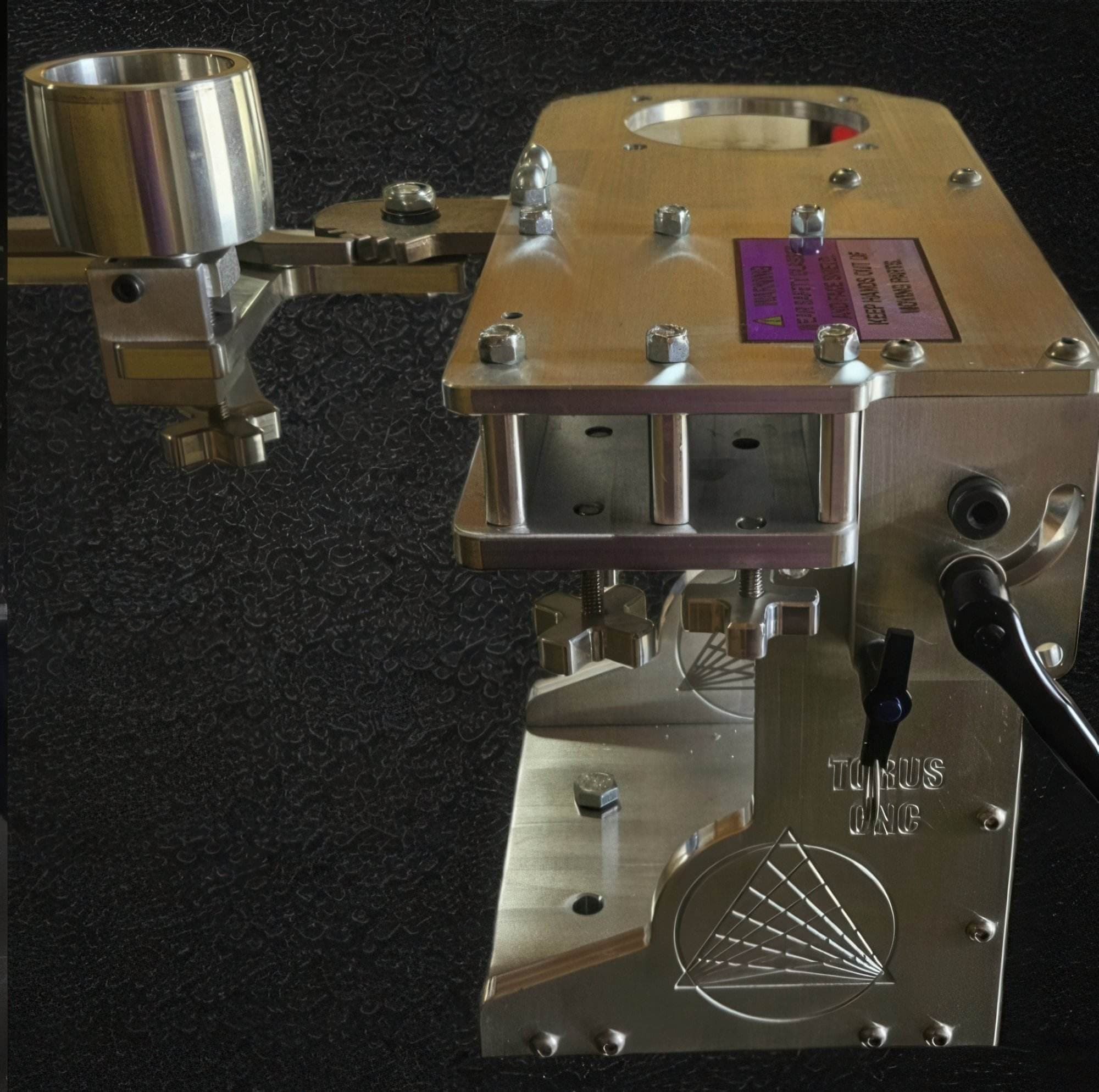 Top-down view of a Torus CNC aluminum grinder chassis with a safety warning label and bolts