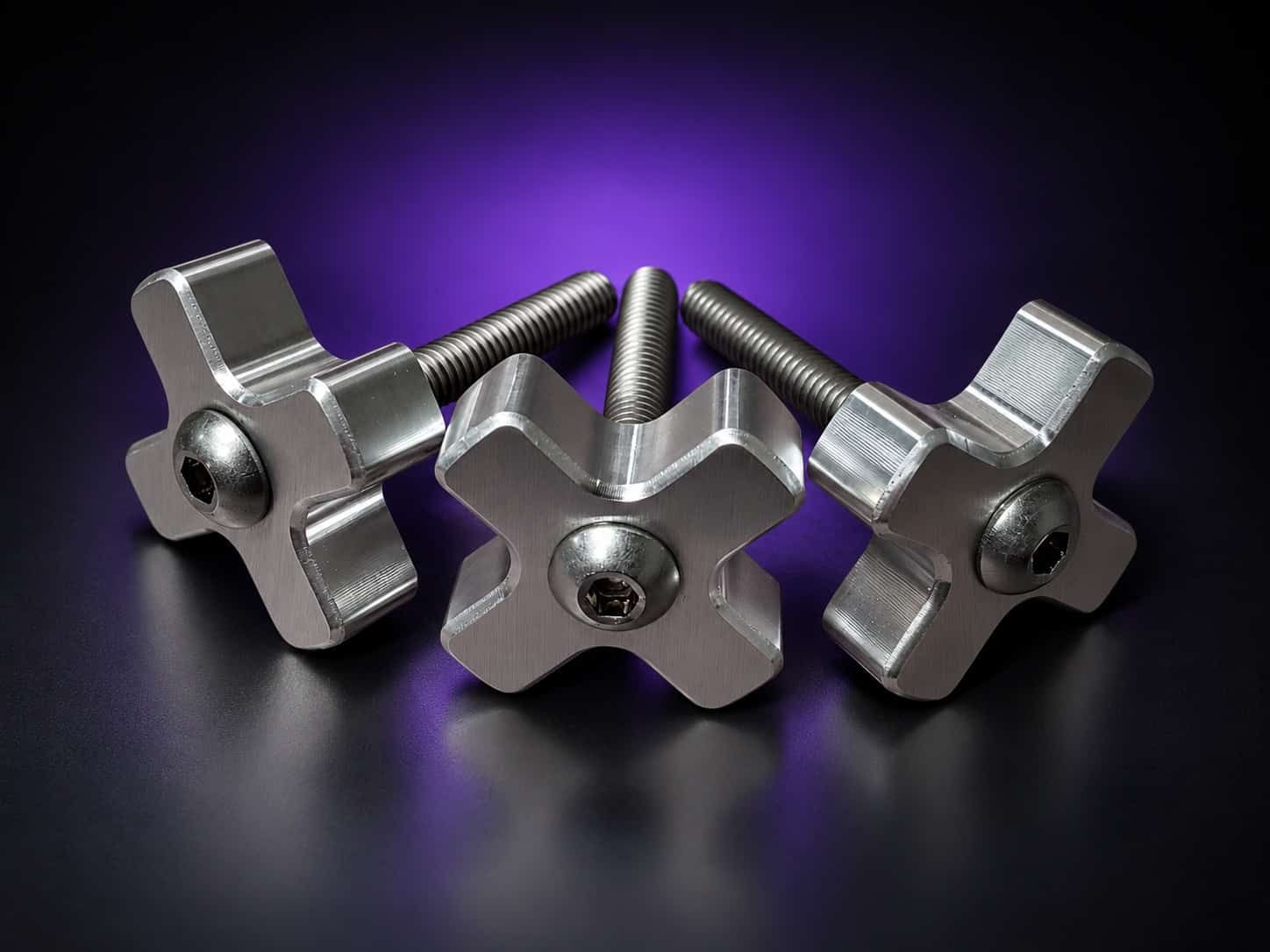 Three aluminum cross-shaped tension knobs with stainless threaded studs, angled view, on black with a purple glow