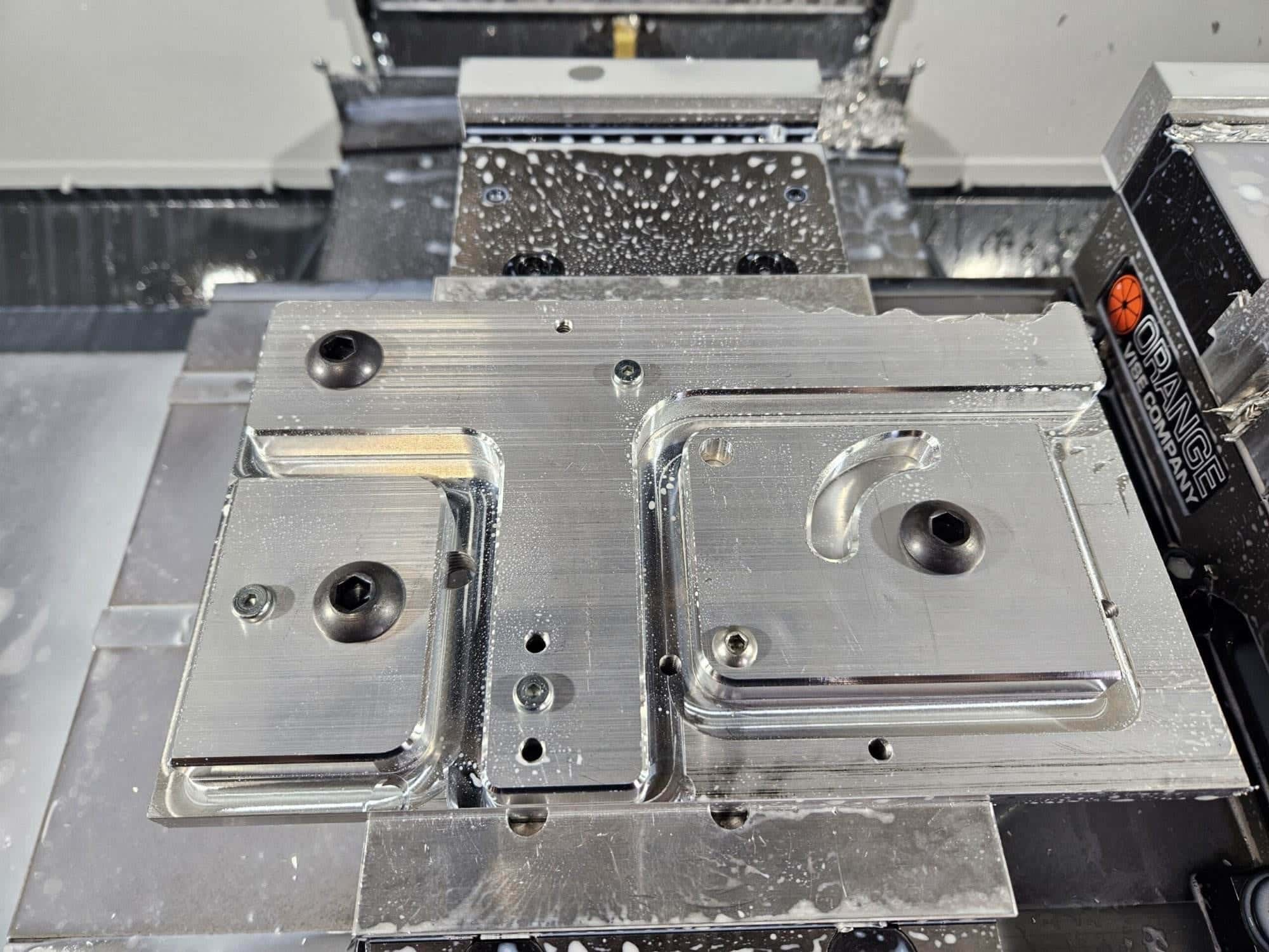 Machined aluminum grinder base plate with milled pockets clamped on the mill table, still wet with coolant