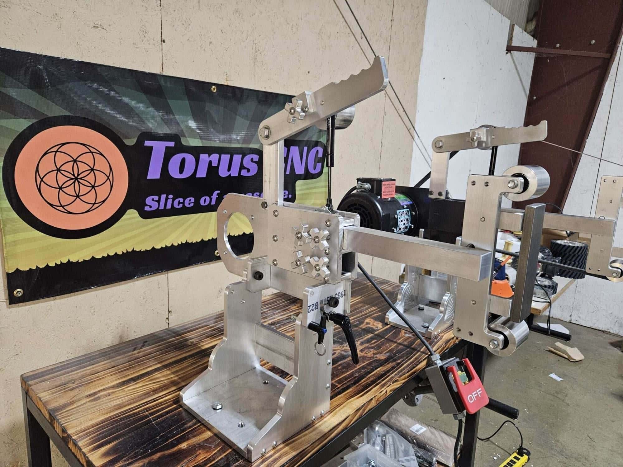 Torus CNC belt grinder assembly displayed in the shop in front of the Torus CNC banner sign