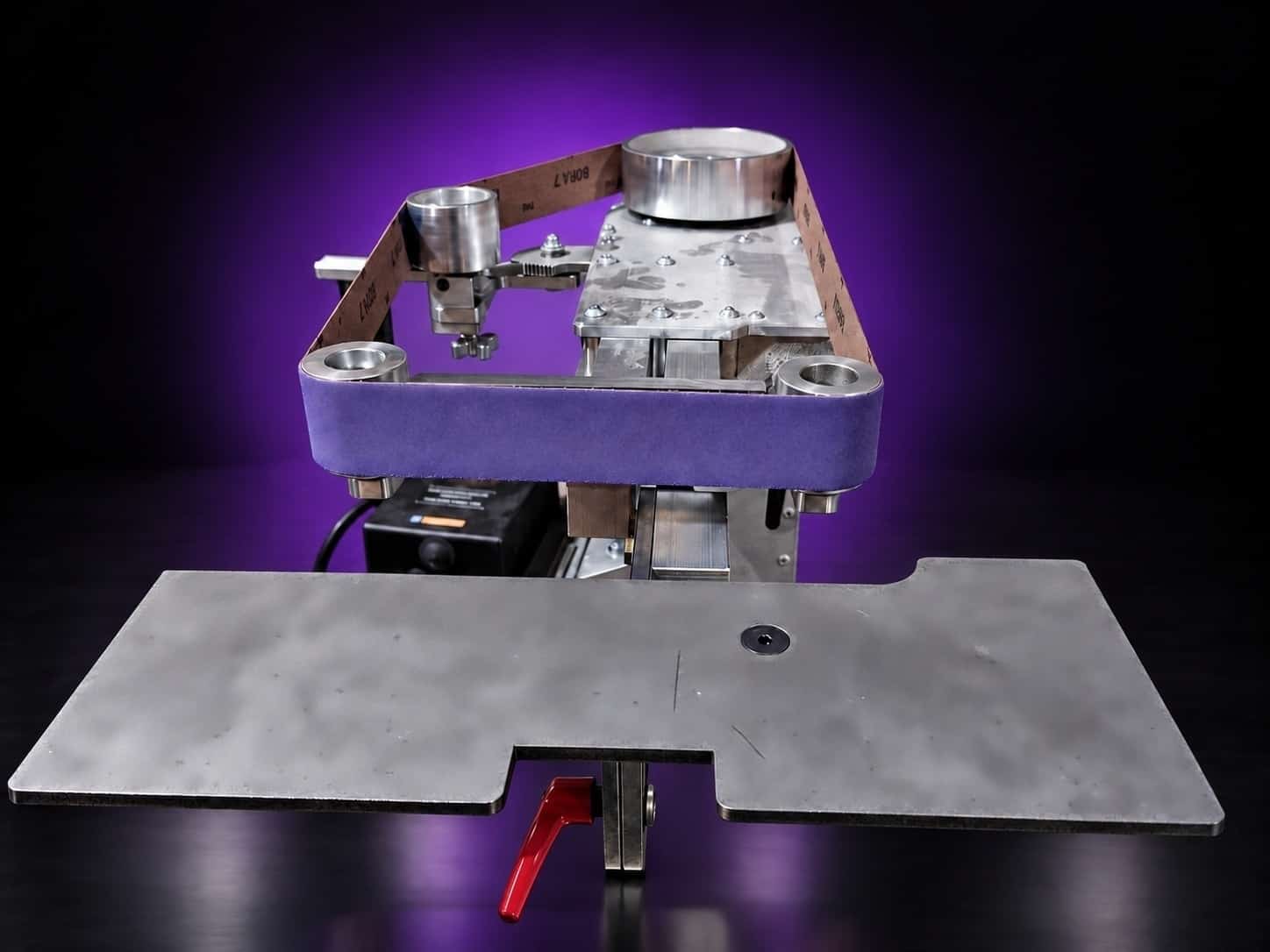 Steel work table in front of a grinder running a purple abrasive belt, seen from the table side, on black with a purple glow