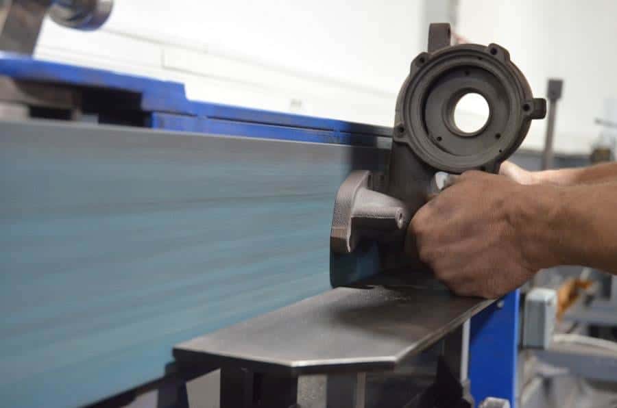Shop worker's hands grinding a cast aluminum housing against a belt grinder platen with dust flying